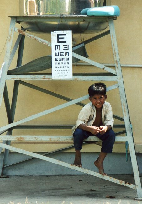 Kambodschanischer Junge hockt auf einem Metallgestell, an dem eine Sehtest-Tafel angebracht ist